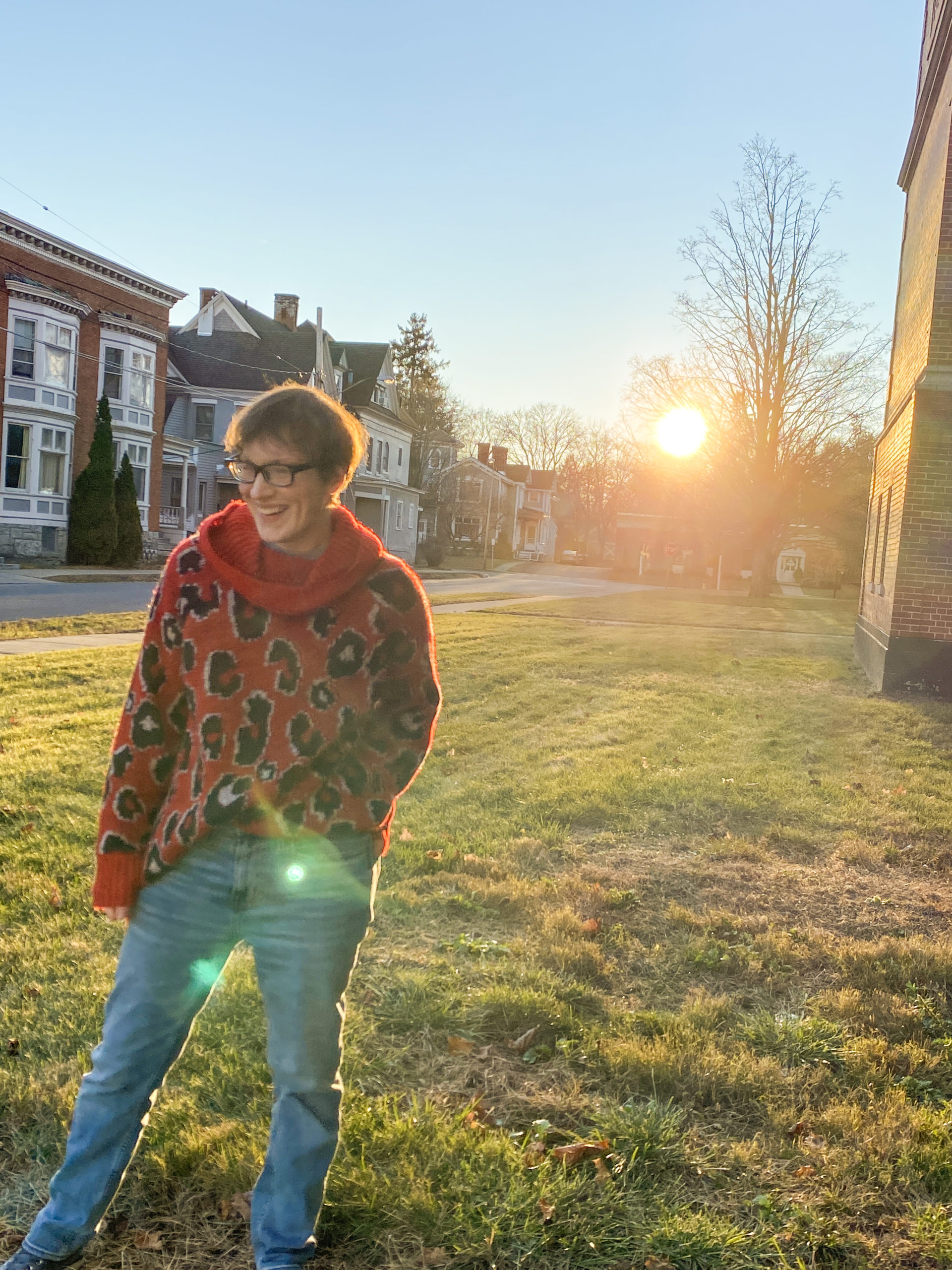 Image of effeminate young white man with a decorative brown scarf around his shoulders and short brown hair. The background is streaks of blue, pink, orange, and yellow lines.